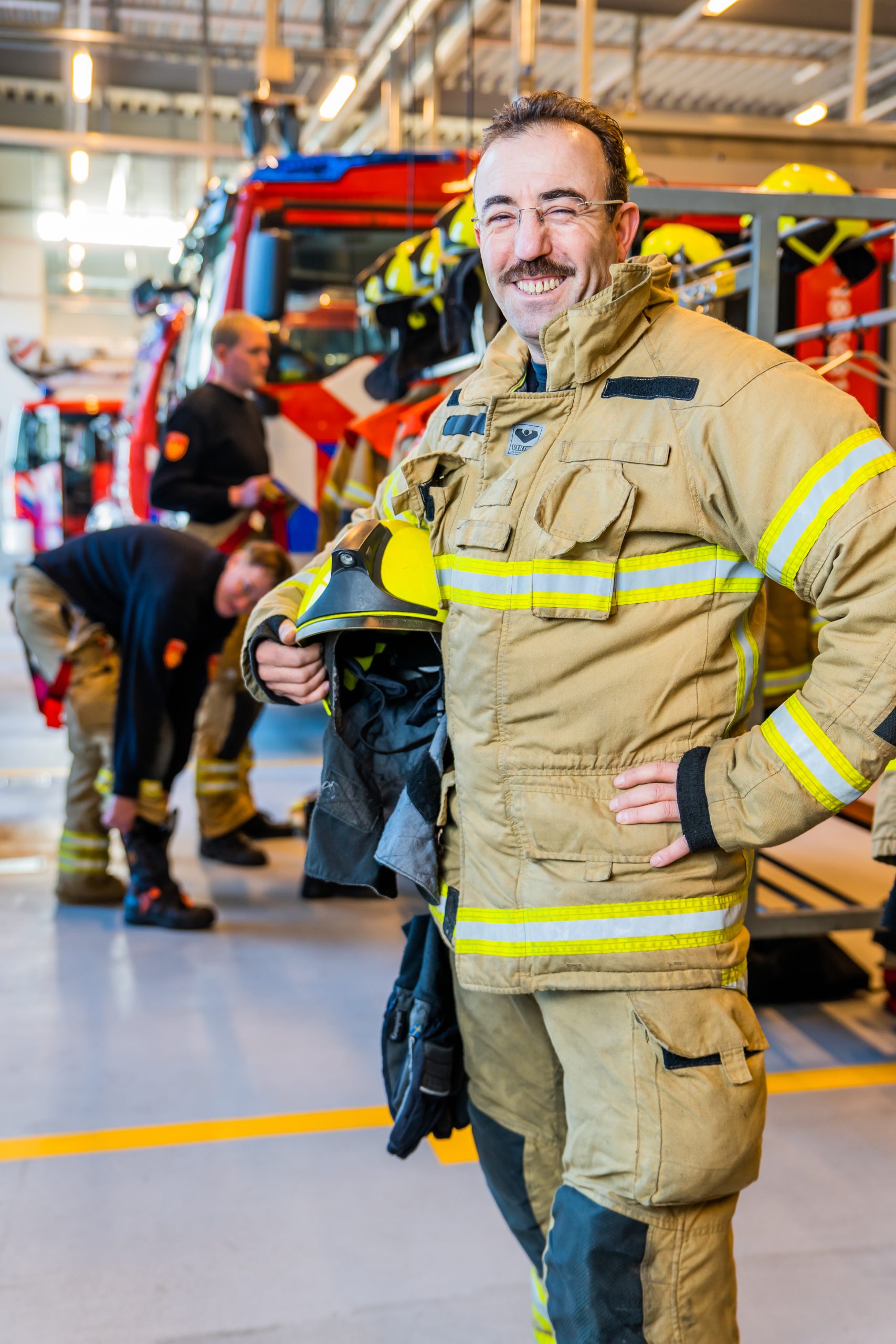 Cemal kijkt in de camera heeft een brandweer outfit aan en een helm in zijn hand. Hij staat in de kazerne.