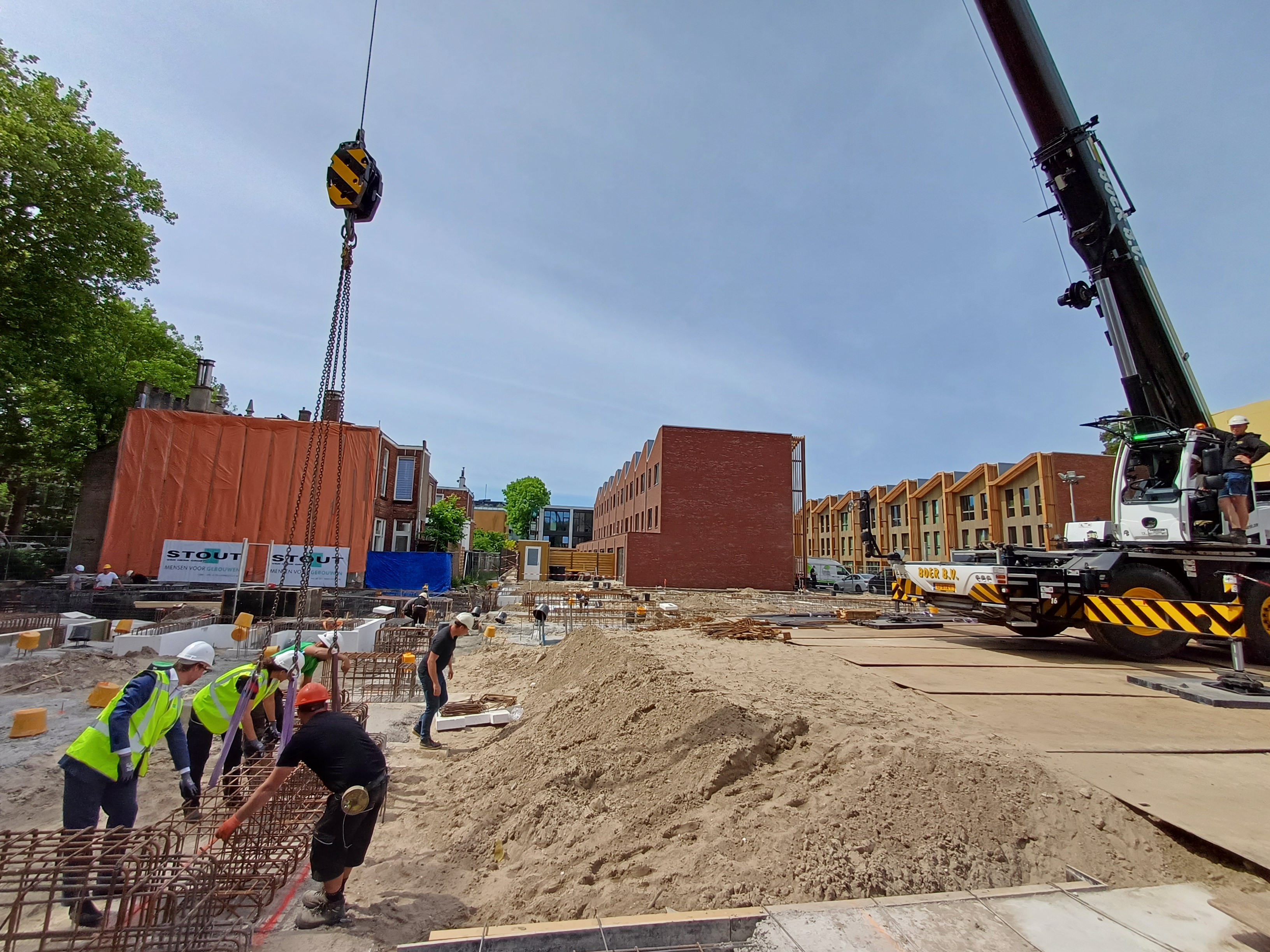Bouwplaats van de nieuwe brandweerkazerne in Dordrecht. Bouwvakkers en vertegenwoordigers van de gemeente, bouwonderneming en Veiligheidsregio plaatsen samen een groot stalen frame in de grond. Rechts staat een grote kraan die helpt bij het tillen van het frame. Op de achtergrond zijn huizen en bouwmaterialen te zien.