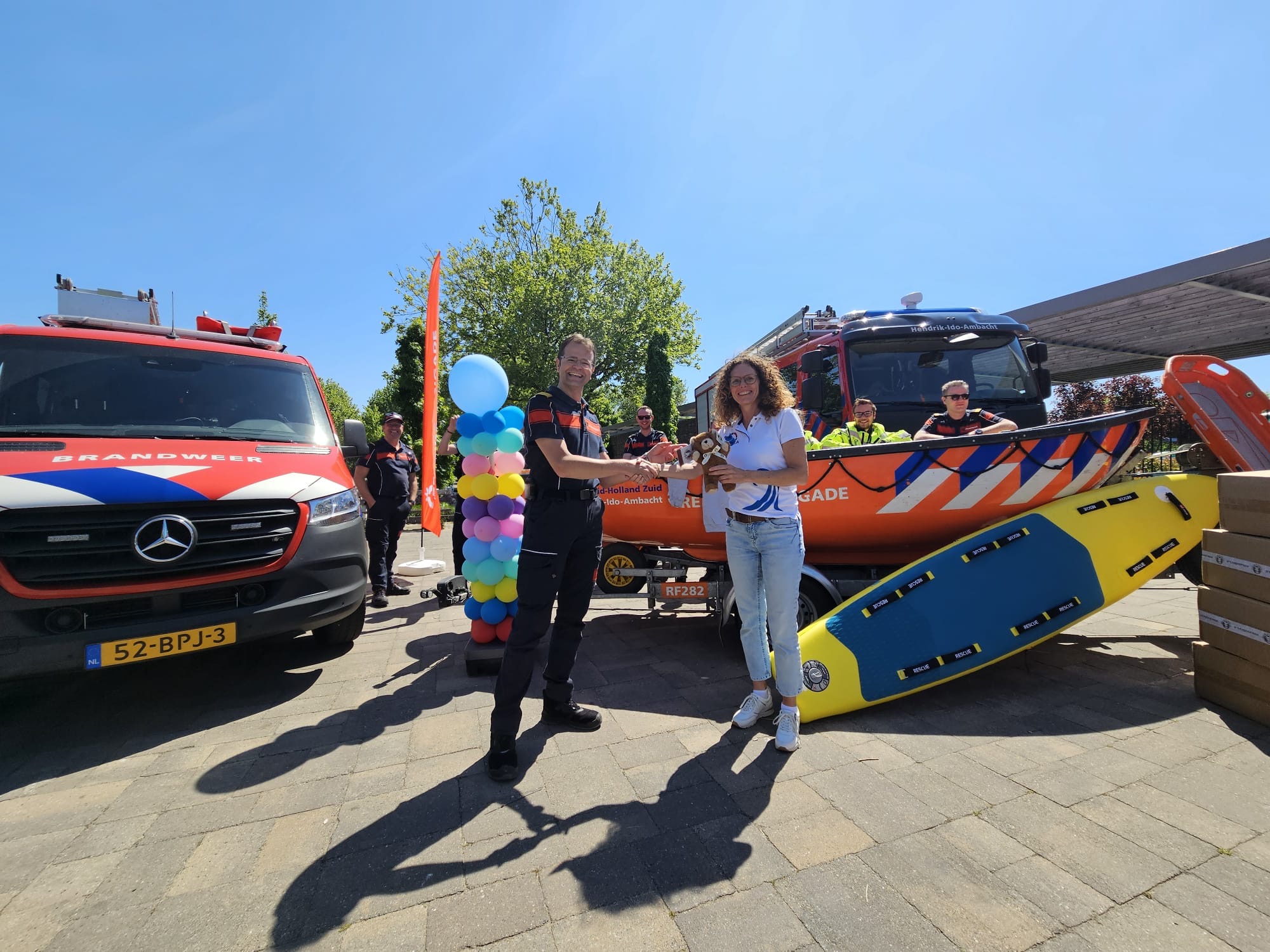 Voorzitter van Reddingsbrigade Hendrik-Ido-Ambacht overhandigt troostbeer aan directeur brandweerzorg Marko Redelijkheid. Op de achtergrond brandweer- en reddingsvoertuigen.
