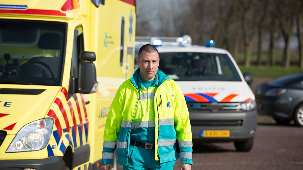 Ambulancemedewerker loopt langs ambulance met politieauto op achtergrond