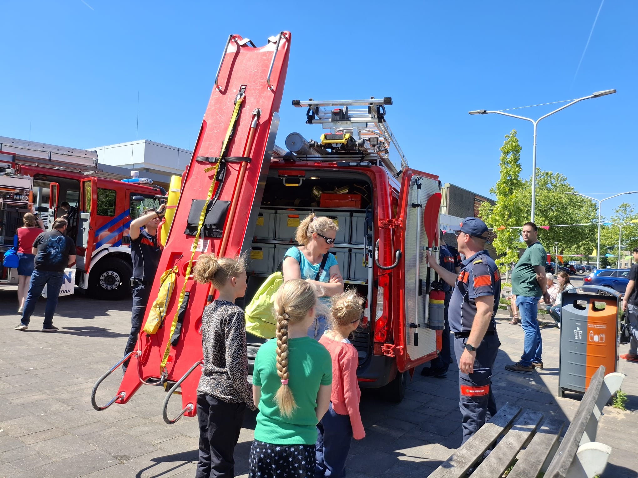 Kinderen en volwassenen bekijken een brandweerwagen van dichtbij. Brandweermensen geven uitleg en tonen reddingsmateriaal, waaronder een grote rode brancard. Op de achtergrond staan meer brandweervoertuigen.