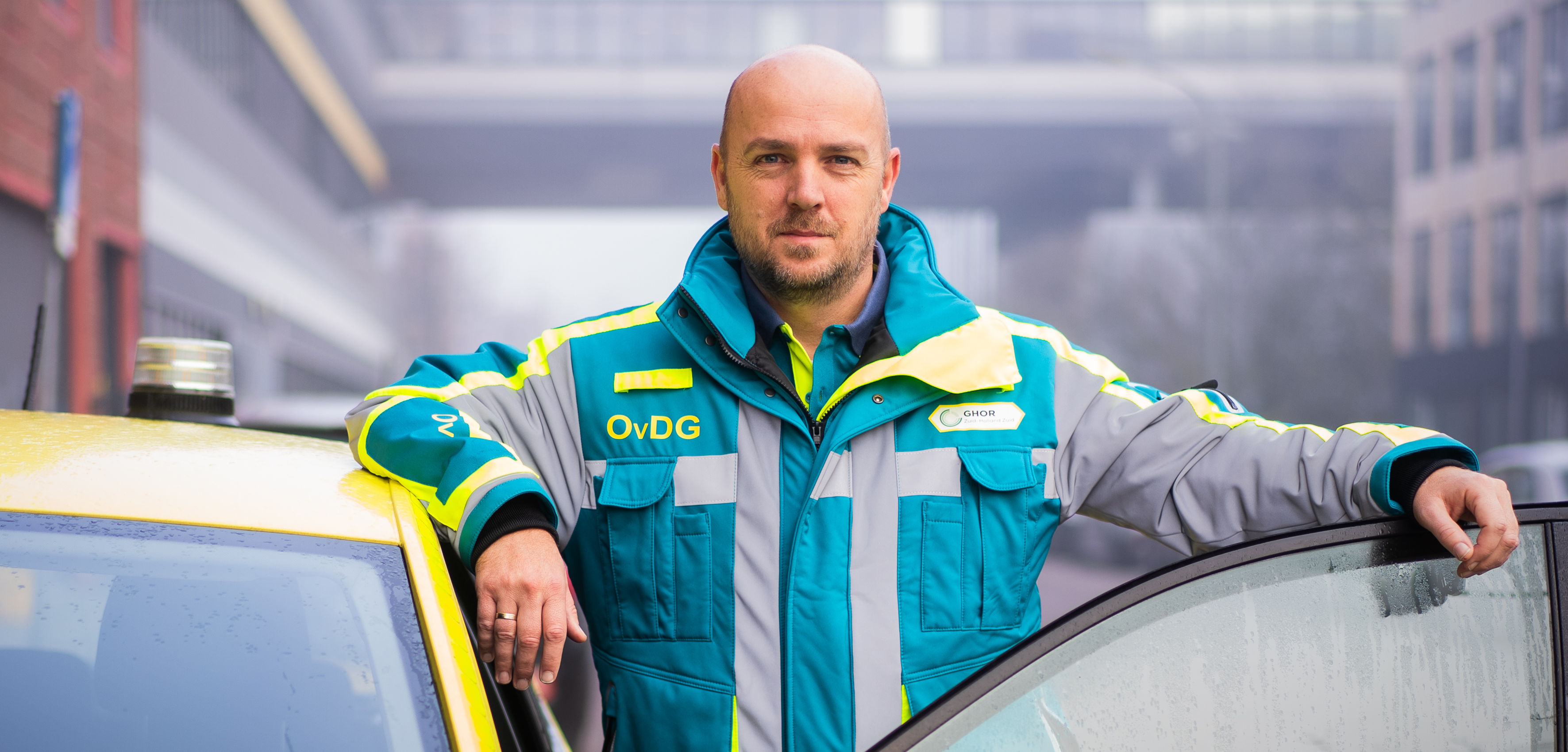 Sander staat in geneeskundige outfit tussen opengeslagen autodeur van ambulanceauto en kijkt recht in de camera