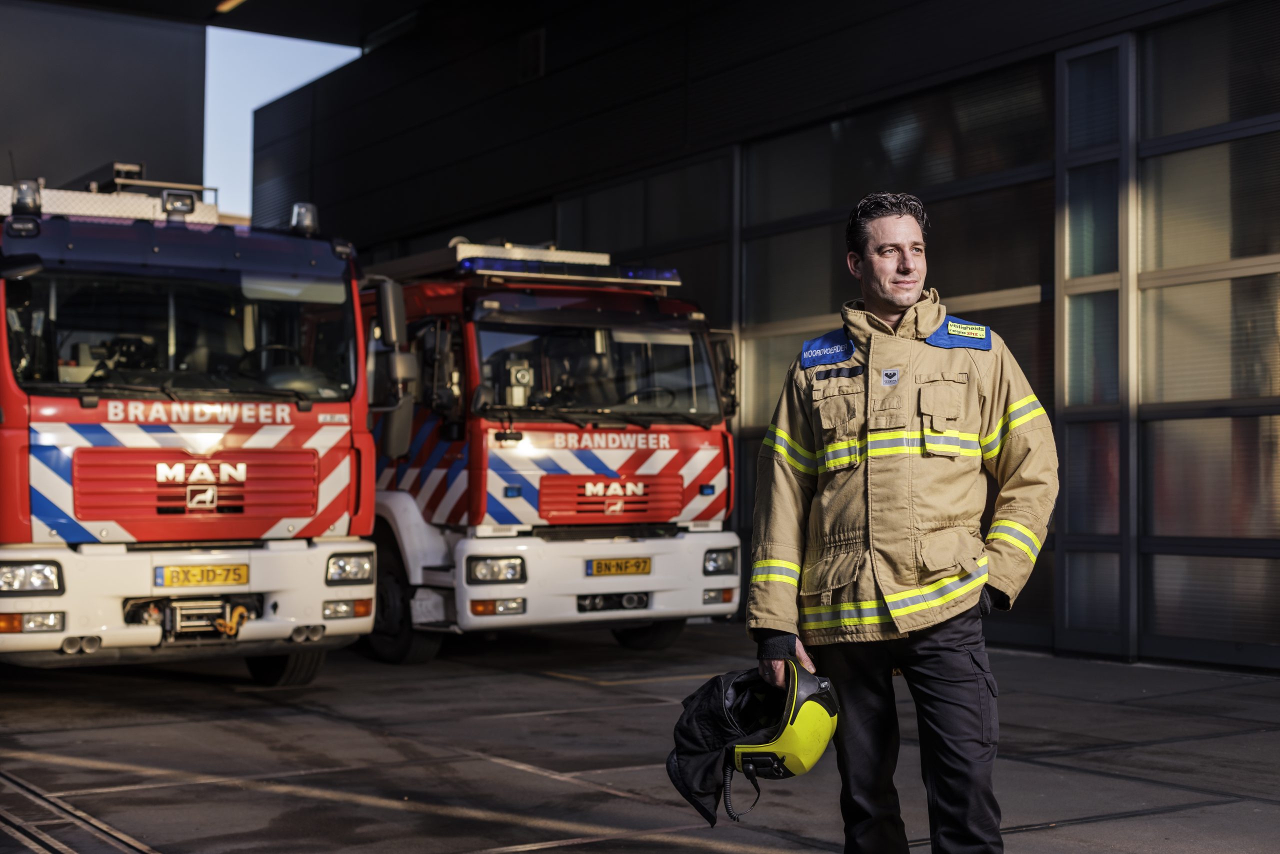 Marc staat voor twee brandweerauto's. Hij heeft zijn werk outfit aan en een helm in zijn hand en zijn andere hand in zijn zak.